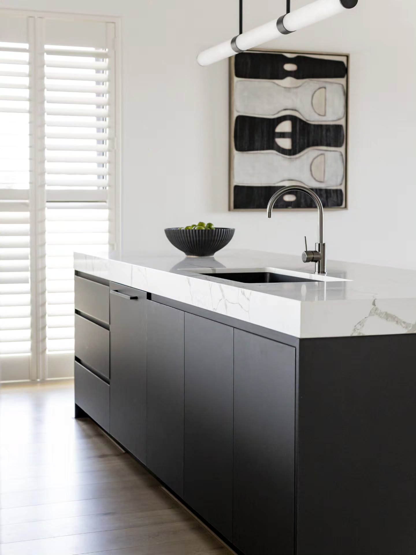 Black fluted island with an integrated sink beneath monochrome artwork and plantation shutters — Monochrome Residence